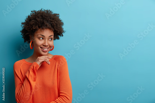 Studio shot of ethnic curly woman touches chin gently, feels happy and delighted, turns face aside, wears bright orange jumper and earrings, isolated over blue background, blank space for your text