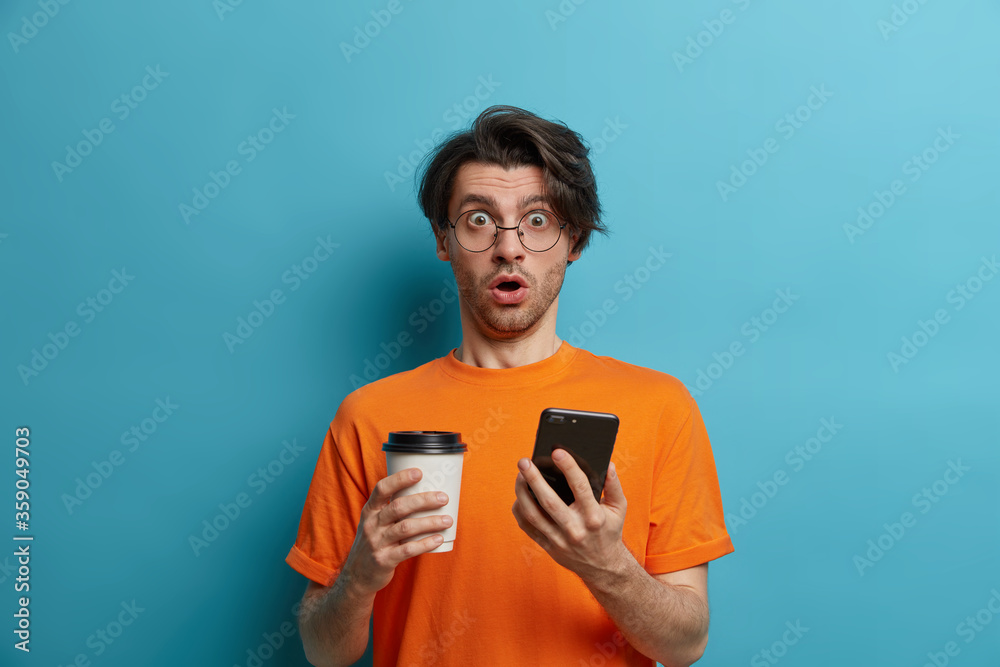 Photo of stunned Caucasian man reads message or astonished impressive article on website via cell phone, holds breath and stares at camera, drinks coffee to go, holds disposable cup, stands indoor