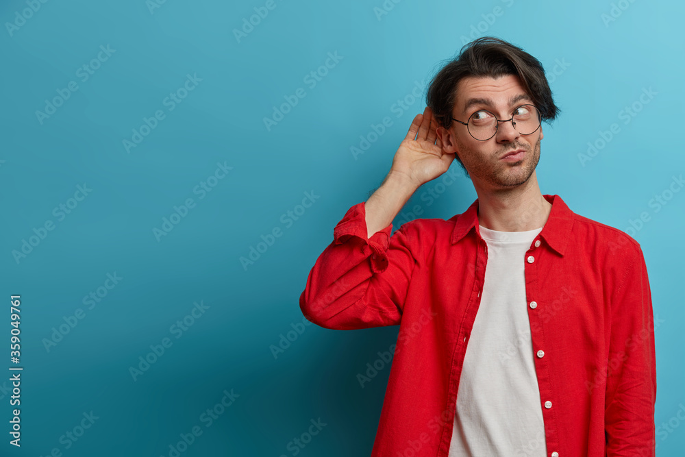 Curious man likes gossips, wants to overhear secret information, wears optical glasses and red shirt, tries to hear womans phone conversation, listens attentively keeps hand near ear for eavesdropping Curious man likes gossips, wants to overhear secret information, wears optical glasses and red shirt, tries to hear womans phone conversation, listens attentively keeps hand near ear for eavesdropping
