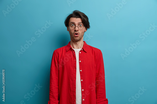 Half length shot of embarrassed shocked hipster guy with trendy hairstyle, gasps from wonder, stunned by unexpected news, wears round spectacles, red stylish shirt, poses indoor over blue background