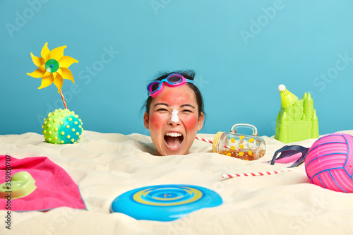 Emotive displeased Asian female model screams loudly, keeps mouth opened, feels tired of staying in sand, sunbathes during sunny day, poses at seashore by water, surrounded with beach accessories. © wayhome.studio 