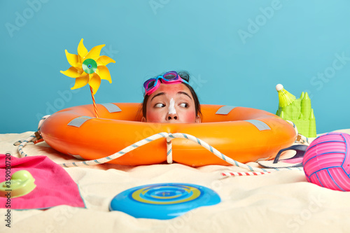 Horizontal shot of woman head sticks out from lifebuoy, looks curiously somewhere at beach, wears swimming goggles, tries to hide from sun, spends time actively at seaside, enjoys hot summer © wayhome.studio 