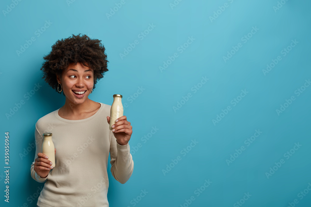 Photo of pleased dark skinned curly girl holds almond milk in glass bottle, finds alternative drink, smiles broadly, looks aside, poses against blue background, empty space for your information