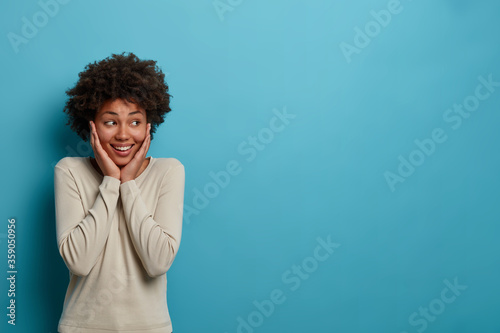 So excited! Cheerful pleasant looking dark skinned woman touches cheeks with both hands, looks aside with toothy smile, has positive attitude to life, isolated on blue background, copy space aside
