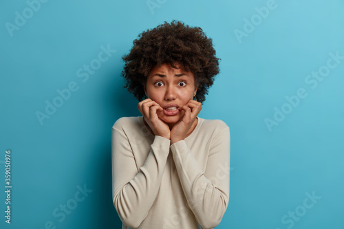 Portrait of puzzled nervous woman grabs face and looks anxiously at camera, trembles from fear, wears casual jumper, isolated on blue background has depression panic attack. People, facial expressions