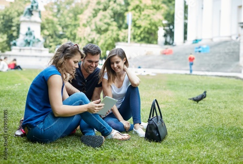 Young friends sitting in park on the grass, using tablet computer. 