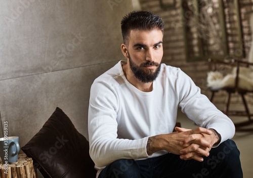 Portrait of trendy bearded man in casual clothes sitting at home, looking at camera. 