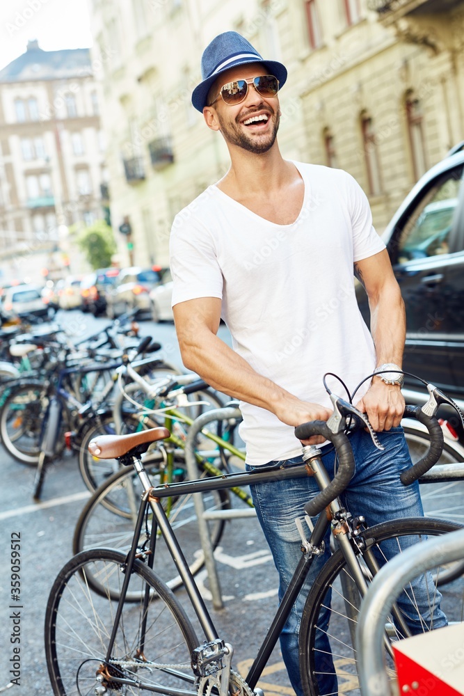 Trendy young man in hat pulling bicycle on street in big city in summer. 