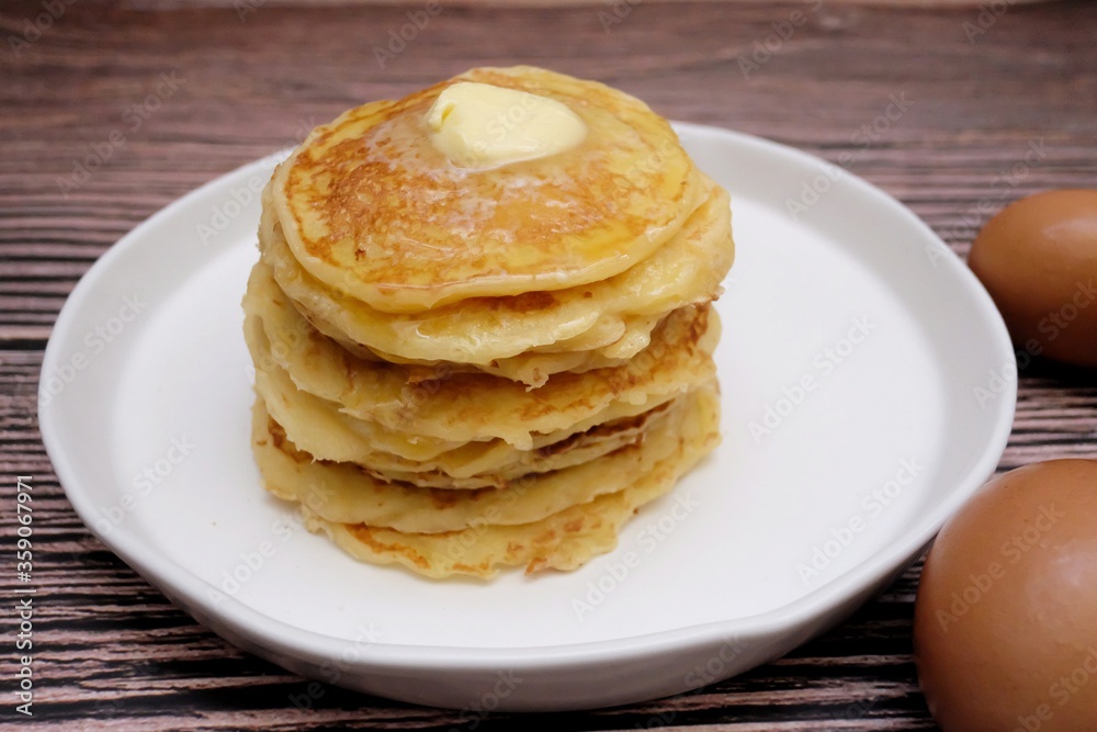 In selective focus butter pancake in a white plate with blurred eggs and old wooden dinning table background In selective focus butter pancake in a white plate with blurred eggs and old wooden dinning table background