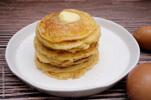 In selective focus butter pancake in a white plate with blurred eggs and old wooden dinning table background