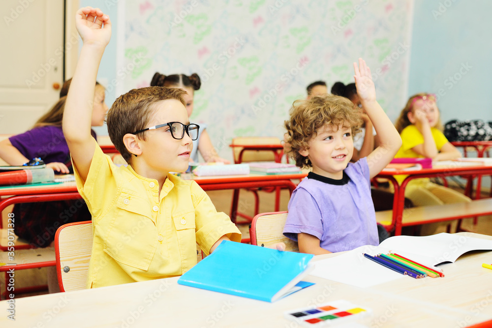 Fototapeta premium two small school children-a boy with curly hair and a boy in a yellow shirt and glasses-smile and hold up their hand against the background of the classroom and a group of classmates. Back to school
