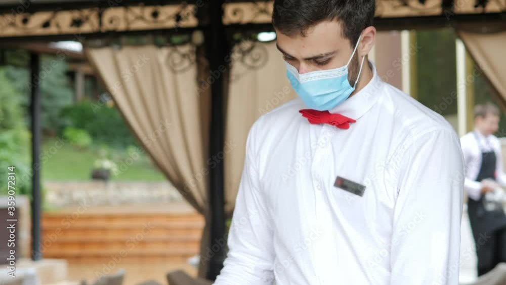 A waiter in a medical protective mask serves the table in the restaurant A waiter in a medical protective mask serves the table in the restaurant