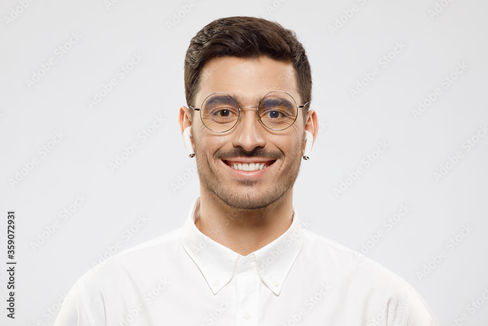 Portrait of young smiling handsome IT specialist dressed in white shirt, wearing eyeglasses and wireless earphones, isolated on gray background Portrait of young smiling handsome IT specialist dressed in white shirt, wearing eyeglasses and wireless earphones, isolated on gray background
