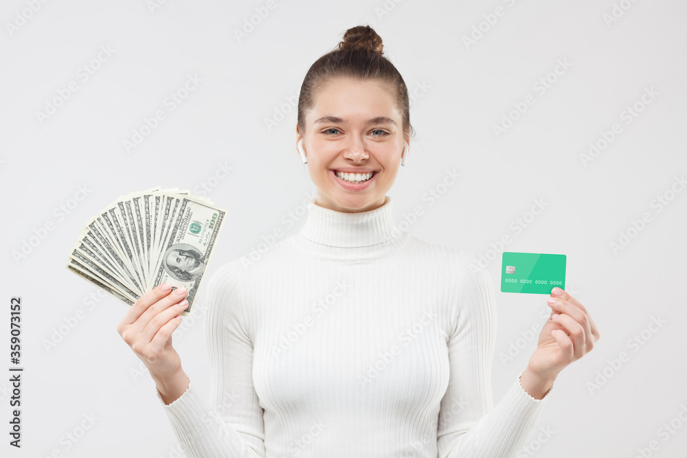 Young female smiling happily while holding fan of hundred dollar banknotes in one hand and credit card in another, satisfied with cash, isolated on gray background Young female smiling happily while holding fan of hundred dollar banknotes in one hand and credit card in another, satisfied with cash, isolated on gray background