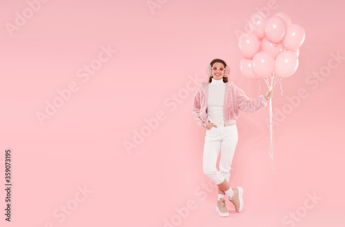 Full length portrait of young happy birthday girl dressed in winter outfit, wearing ear warmers and glasses, holding balloons, isolated on pink background with copy space