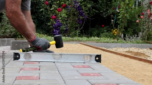 A gloved craftsman is laying concrete tiles using a bubble level and a rubber mallet. Tile laying sounds. Brick walkway paved with professional paver. Laying gray concrete paving slabs in the courtyar