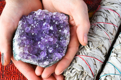 A close up image of a hand holding beautiful purple amethyst cluster next to two white sage bundles. 