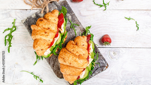 Fresh croissant sandwich strawberrieswith arugula, brie cheese, camembert. Delicious breakfast or snack on a light background, top view