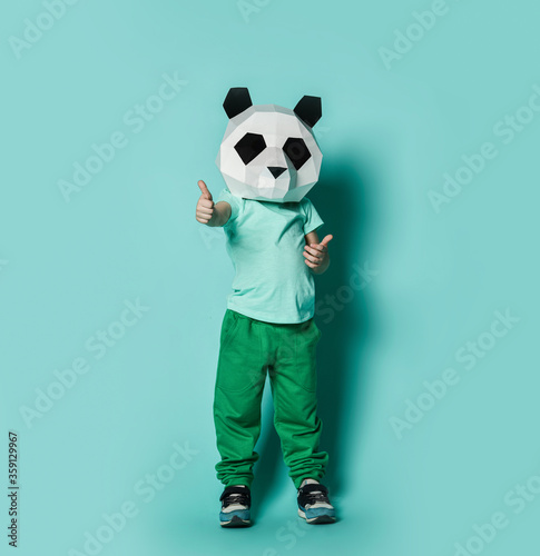 Fototapeta Naklejka Na Ścianę i Meble -  Kid boy in panda mask white blank t-shirt and green pants stands gesturing thumbs up sign with both hands