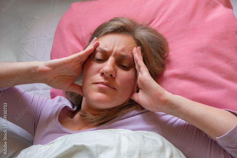 Middle aged woman has a headache. 40 years women lying in the bed and touches the head with hands. She is unwell and sick. Girl close-up portrait with health problems Middle aged woman has a headache. 40 years women lying in the bed and touches the head with hands. She is unwell and sick. Girl close-up portrait with health problems