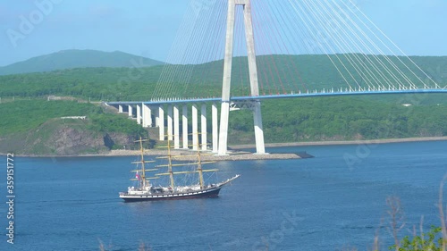 A beautiful three-masted sailboat passes under the Russian bridge in Vladivostok against the backdrop of the Russian island.