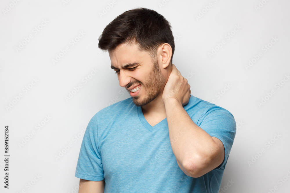 Young handsome man in blue tshirt, touching his neck because it hurts after long working hours, feeling exhausted and worried, isolated on gray background Young handsome man in blue tshirt, touching his neck because it hurts after long working hours, feeling exhausted and worried, isolated on gray background