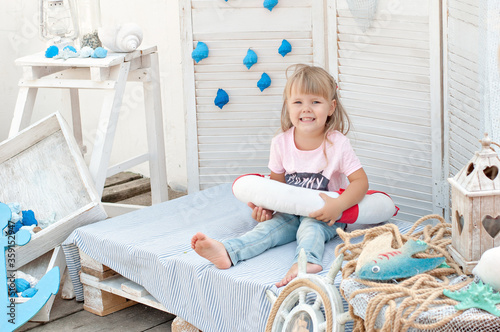 Beautiful little blonde girl with blue eyes looks at the camera and laughs. The baby plays with a lifebuoy and shells on the background of marine decoration. Sea adventures and games for children. 