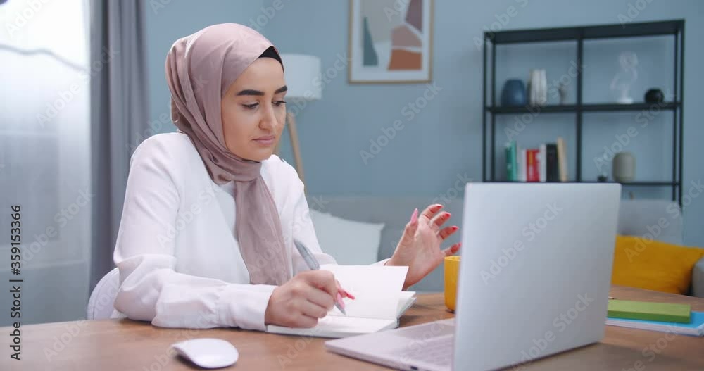 Muslim student studying at desk in room, using laptop and notebooks. Girl wearing hijab preparing for exams. Female enjoying hot tea from cozy yellow cup. Student lifestyle, modern islam concept Muslim student studying at desk in room, using laptop and notebooks. Girl wearing hijab preparing for exams. Female enjoying hot tea from cozy yellow cup. Student lifestyle, modern islam concept