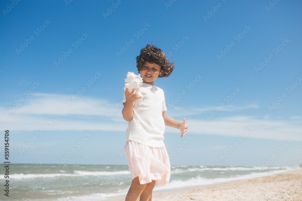 Little girl wearing light t-short and light pink skirt at the sea beach with a big beautiful seashell. Summer vacation Little girl wearing light t-short and light pink skirt at the sea beach with a big beautiful seashell. Summer vacation