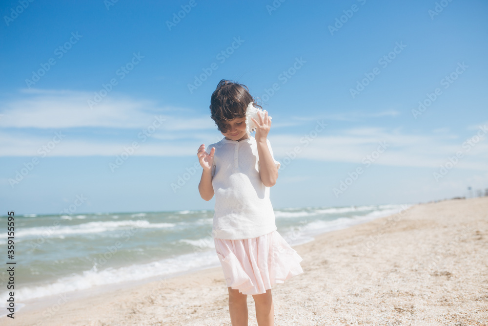 Little girl wearing light t-short and light pink skirt at the sea beach with a big beautiful seashell. Summer vacation Little girl wearing light t-short and light pink skirt at the sea beach with a big beautiful seashell. Summer vacation