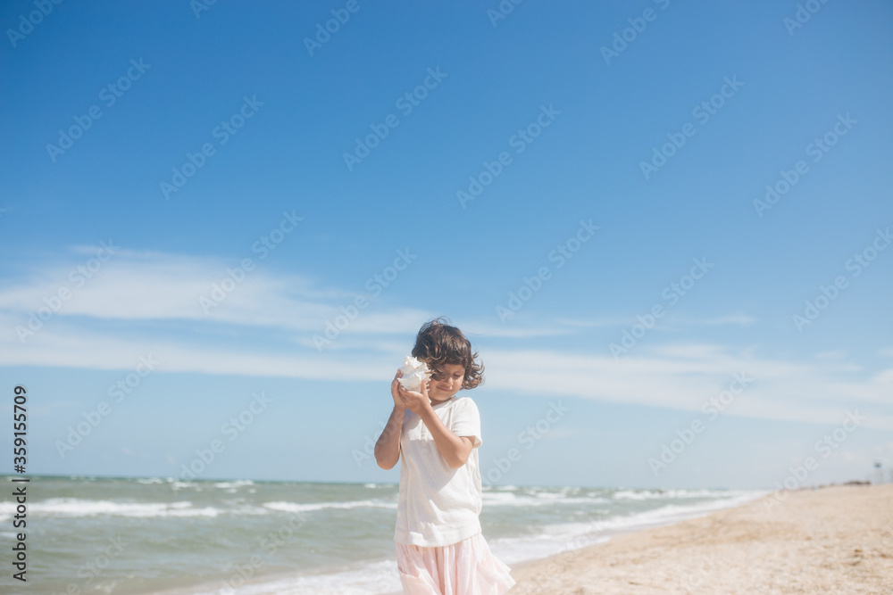 Little girl wearing light t-short and light pink skirt at the sea beach with a big beautiful seashell. Summer vacation Little girl wearing light t-short and light pink skirt at the sea beach with a big beautiful seashell. Summer vacation