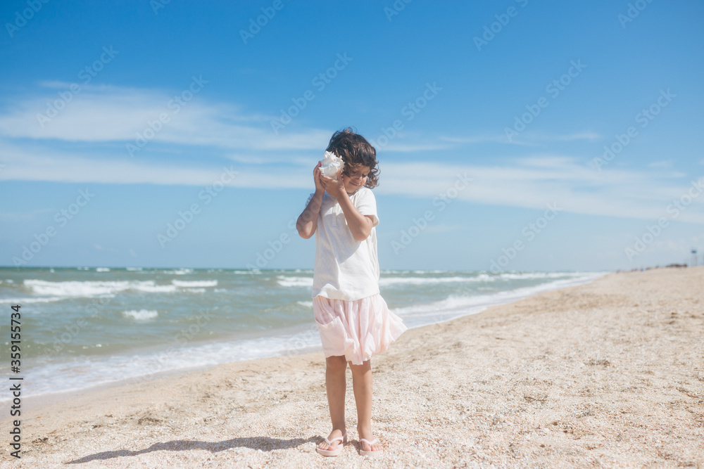 Little girl wearing light t-short and light pink skirt at the sea beach with a big beautiful seashell. Summer vacation