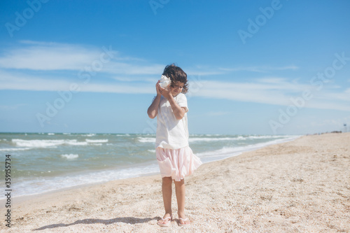 Little girl wearing light t-short and light pink skirt at the sea beach with a big beautiful seashell. Summer vacation © sweetlaniko