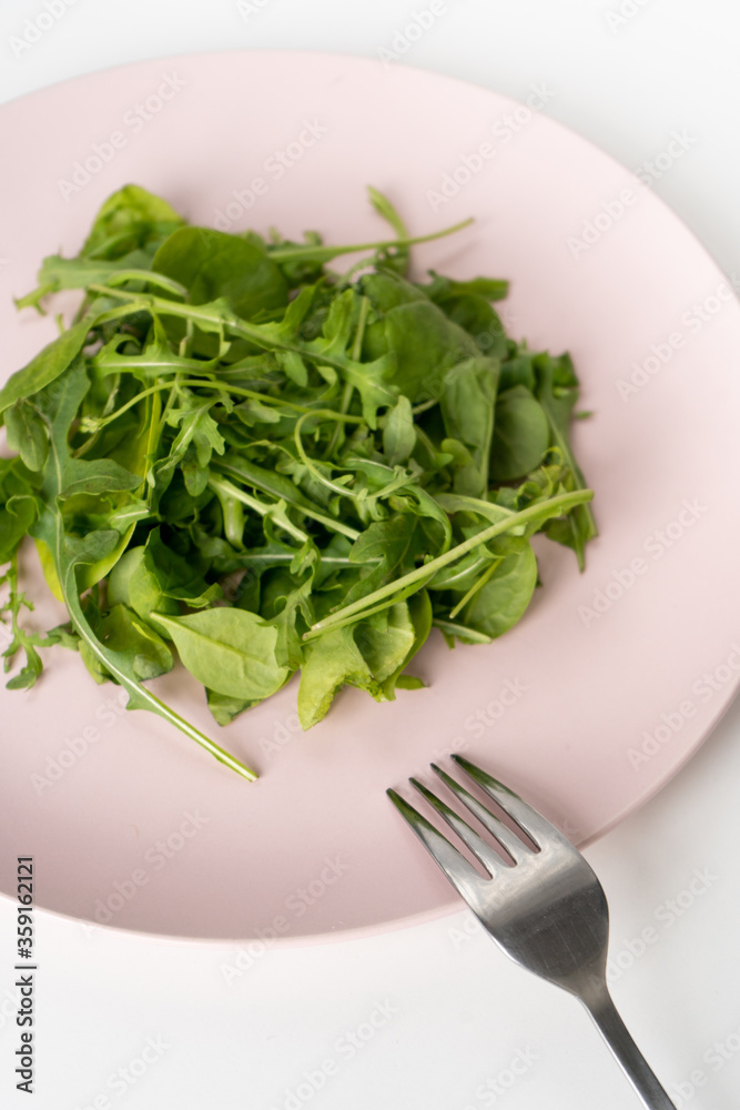 Fresh spinach and arugula leaves on a pink plate and a fork on a white table. Dietary green salad for a healthy diet. Vertical. Close-up Fresh spinach and arugula leaves on a pink plate and a fork on a white table. Dietary green salad for a healthy diet. Vertical. Close-up