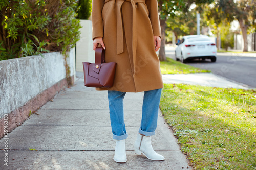Detail of fashionable young woman wearing beige coat, blue jeans and white cowboy boots. She is holding a brown bag. Stylish. Street style.