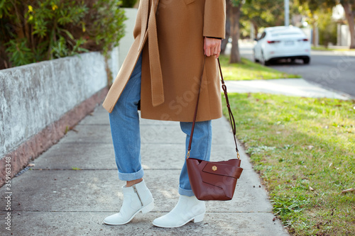 Detail of fashionable young woman wearing beige coat, blue jeans and white cowboy boots. She is holding a brown bag. Stylish. Street style.