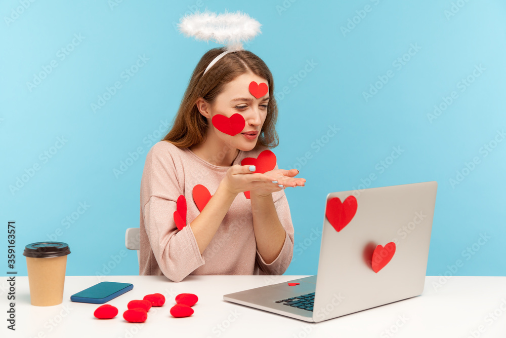 Beautiful angelic woman with kind expression sitting covered with sticker hearts, sending romantic sensual air kiss to laptop screen, dating on video call with lover. indoor studio shot, isolated Beautiful angelic woman with kind expression sitting covered with sticker hearts, sending romantic sensual air kiss to laptop screen, dating on video call with lover. indoor studio shot, isolated