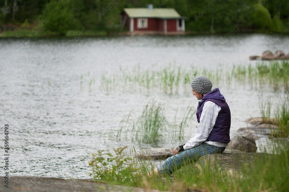 Woman in hat and purple waistcoat relaxing on lakeside in breezy summer day. Woman is spending time alone to find harmony and balance. New vacation approach - responsible travel. Solo travell concept.