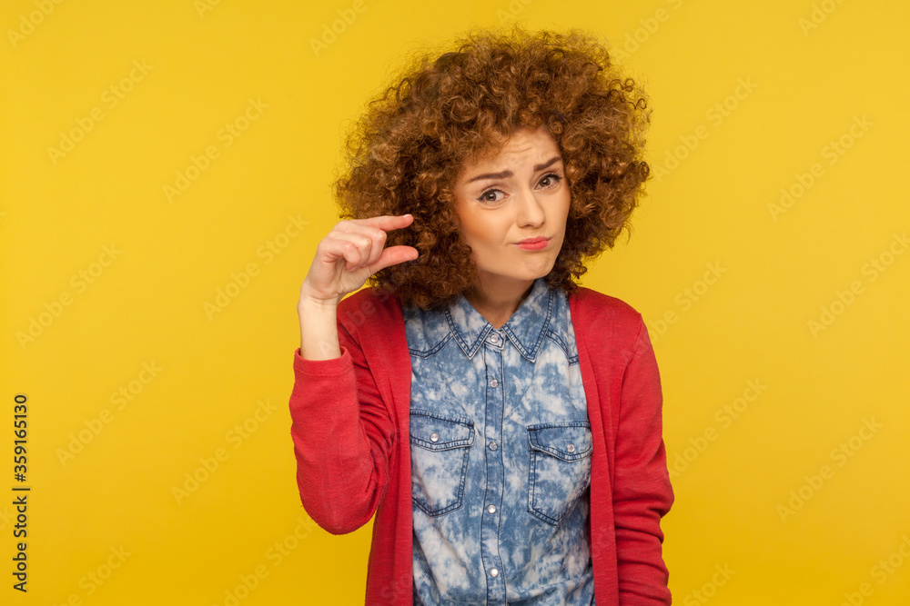 Minimum amount. Portrait of disappointed woman with curly hair showing a little bit gesture, sceptic about centimeter inch size, measuring small scale. indoor studio shot isolated on yellow background