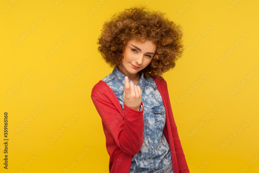 Come here, follow me! Portrait of pretty curly-haired woman looking playfully and calling with one finger, making beckoning gesture, inviting to come. indoor studio shot isolated on yellow background
