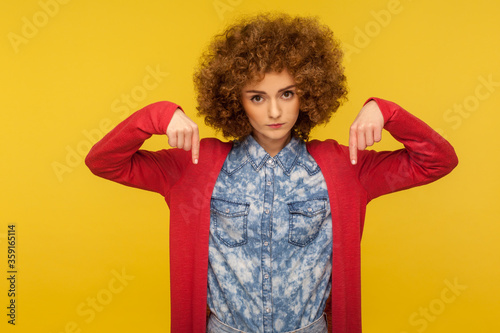 You must start right now! Portrait of angry bossy woman with curly hair pointing finger down, giving order with demanding expression, control concept. indoor studio shot isolated on yellow background © khosrork