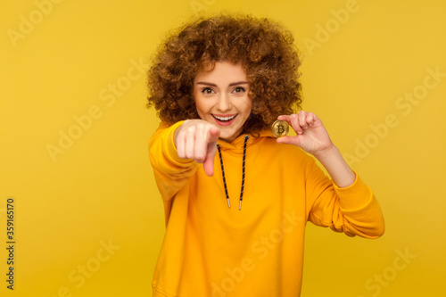 Hey you, buy digital money! Portrait of happy excited curly-haired young woman in urban style hoodie holding golden bitcoin and pointing to camera. indoor studio shot isolated on yellow background