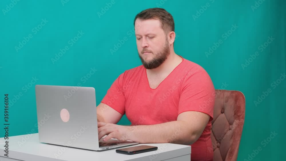 Young happy businessman in a pink t-shirt works on a laptop on a green screen. Portrait of a male talking man looking into his laptop. Man working at his desk in the office. Young happy businessman in a pink t-shirt works on a laptop on a green screen. Portrait of a male talking man looking into his laptop. Man working at his desk in the office.