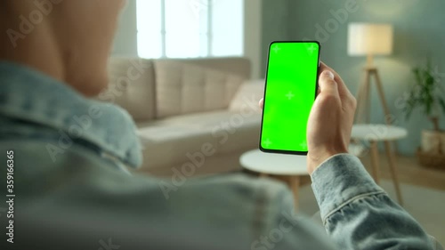 Back View of Young Man at Home Sitting on a Chair using With Green Mock-up Screen Smartphone. Boy is Watcing Content Without Touching Gadget Screen. Modent Technology And Information Concept. photo