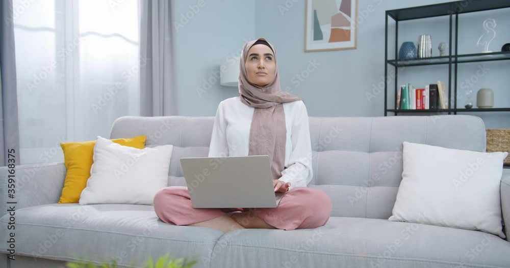 Middle plan of smart young muslim student in hijab working with computer, searching in Internet, browsing, looking thoughtfully into the left. The girl thinking, dreaming, meditating, lost in thought. Middle plan of smart young muslim student in hijab working with computer, searching in Internet, browsing, looking thoughtfully into the left. The girl thinking, dreaming, meditating, lost in thought.