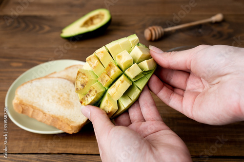 Fototapeta Naklejka Na Ścianę i Meble -  Cutted avocado in female hands, woman cooking toasts with avocado breakfast in the morning on brown wooden table. Close-up. 