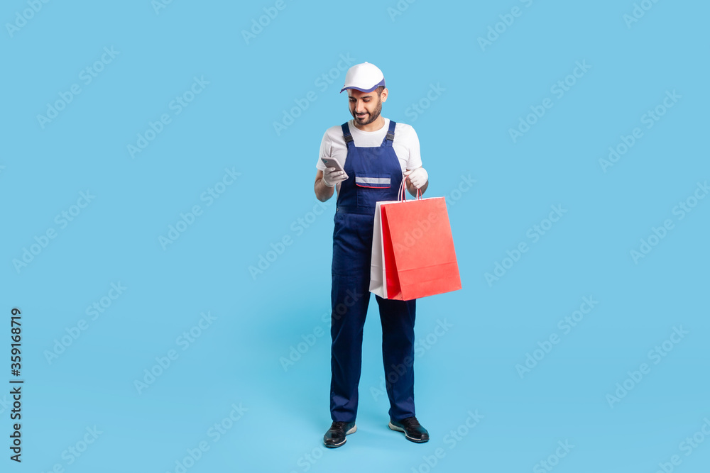 Full length portrait of cheerful courier in workwear holding shopping bags and chatting with client on cell phone, reading message, using mobile app to accept online order at fashion store. isolated Full length portrait of cheerful courier in workwear holding shopping bags and chatting with client on cell phone, reading message, using mobile app to accept online order at fashion store. isolated