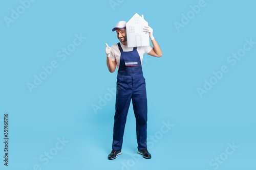 Full length happy labourer in uniform holding paper house on shoulder and showing thumbs up. Mover guy recommending help in relocation, loader services. indoor studio shot isolated on blue background