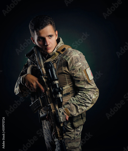 a brutal guy in military airsoft overalls poses with a weapon in his hands on a dark background © rotozey