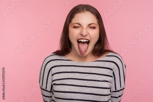Portrait of disobedient funny woman in striped sweatshirt showing tongue out, expressing displeasure, teasing with derisive goofy grimace, naughty behavior. studio shot isolated on pink background